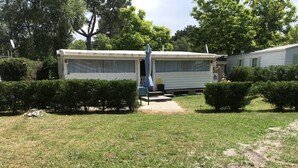 Ein Wohnmobil mit überdachter Veranda und kleiner Terrasse, umgeben von gepflegten Büschen und Bäumen.