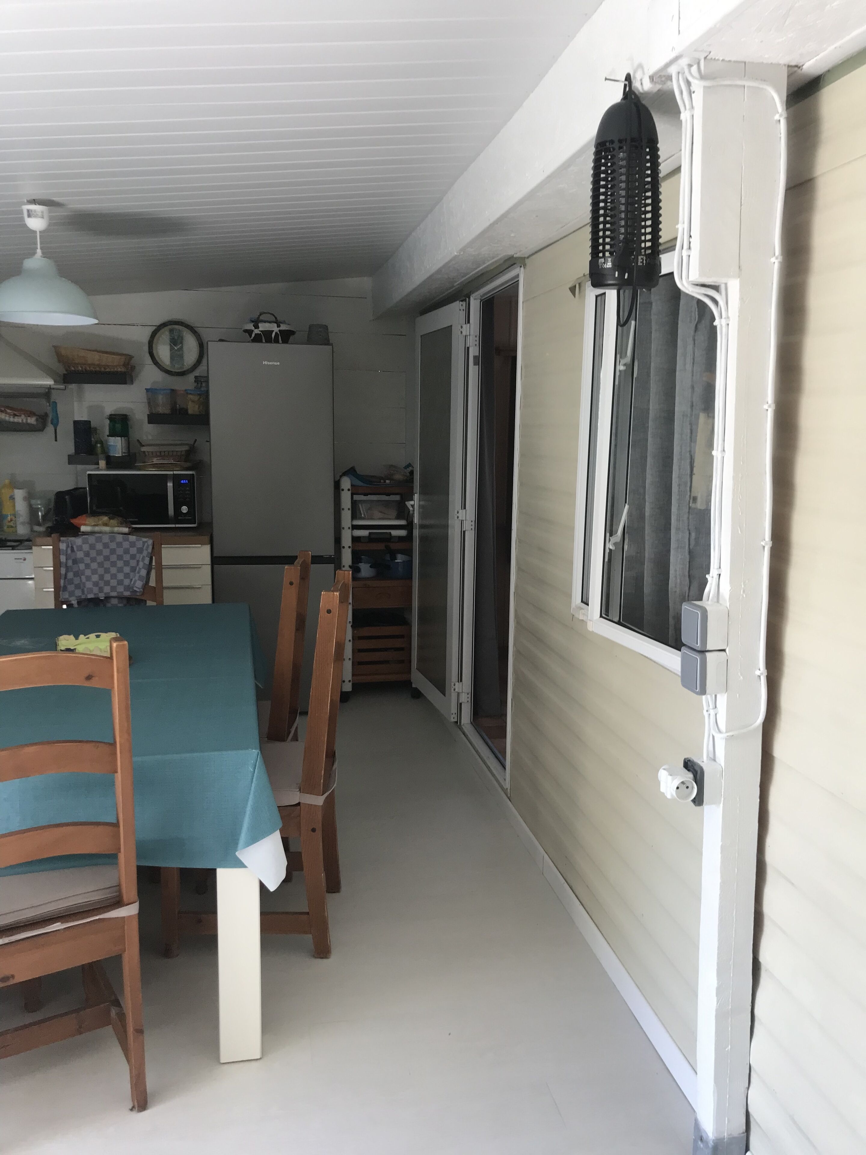 Cocina pequeña con microondas, refrigerador y armarios de madera. Hay una mesa de comedor con sillas y una ventana con persianas.
