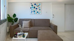 Una sala de estar moderna con un sofá seccional marrón, una mesa de centro blanca con libros y botellas, una gran obra de arte abstracto en la pared y una planta verde grande.