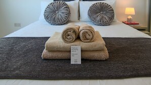 Una cama bien hecha con toallas enrolladas y una tarjeta de agradecimiento, una lámpara en la mesita de noche y un enchufe de pared.