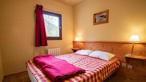 A cozy room with a wooden bed, a window with red curtains, a nightstand with a lamp, and a framed picture on the wall.