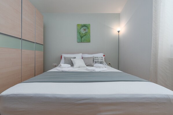 A neatly made bed with pillows and a folded towel, a green floral painting on the wall, and a bedside lamp.