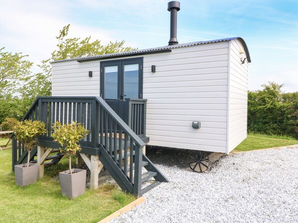 A small, single-story mobile home with a dark wooden staircase leading to a door, surrounded by gravel and grass. - Cwt Y Rhos (Holyhead)