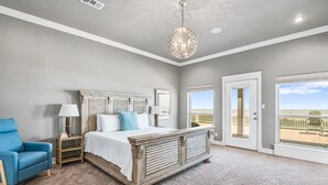 A bedroom with a bed, a blue chair, a nightstand, a lamp, a chandelier, and a view of the beach through a door.