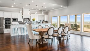 A spacious kitchen with white cabinets, a wooden dining table, and a view of the beach through large windows.