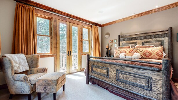 A rustic bedroom with a wooden bed, a chair, and large windows with curtains. - Lodestar #2001 5 Bedroom Home by RedAwning (Mammoth Lakes)