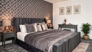 Quarto moderno com parede estampada em padrão geométrico, cama grande com várias almofadas, luminárias de cabeceira, escrivaninha com uma planta e obras de arte emolduradas na parede.