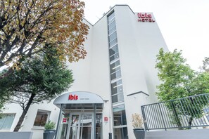 Un hôtel moderne, reconnaissable à son grand panneau « ibis », à son entrée en verre et à son immeuble blanc imposant, surmonté d’une enseigne rouge.