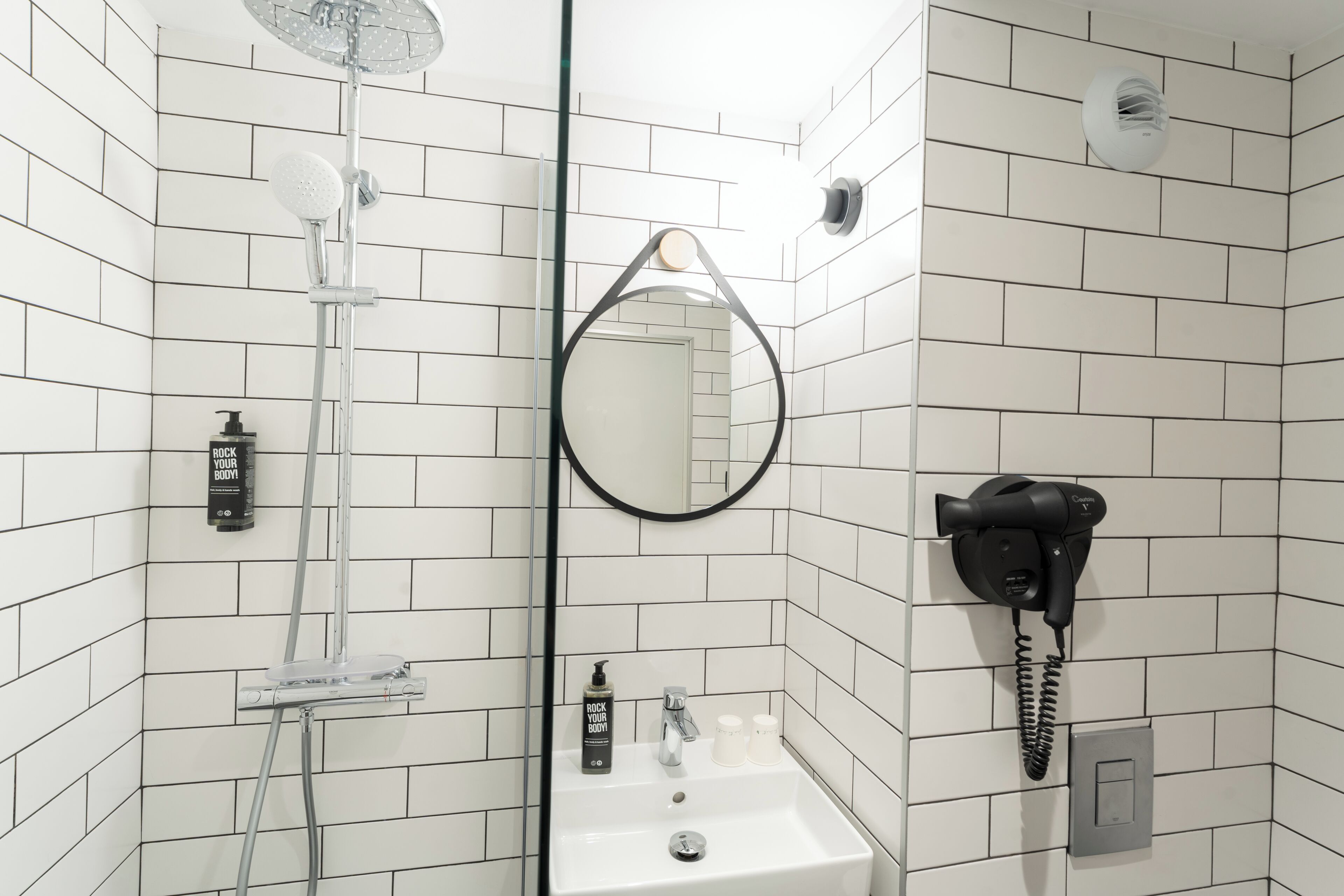 Baño con paredes de azulejos blancos, ducha con cerramiento de vidrio, lavabo con espejo, secador de pelo y dispensador de jabón.