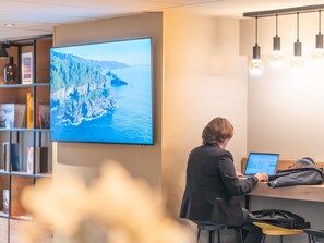 Une personne travaille à un bureau, devant un ordinateur portable. Un grand écran de télévision affiche un paysage côtier, et une bibliothèque contient divers objets.