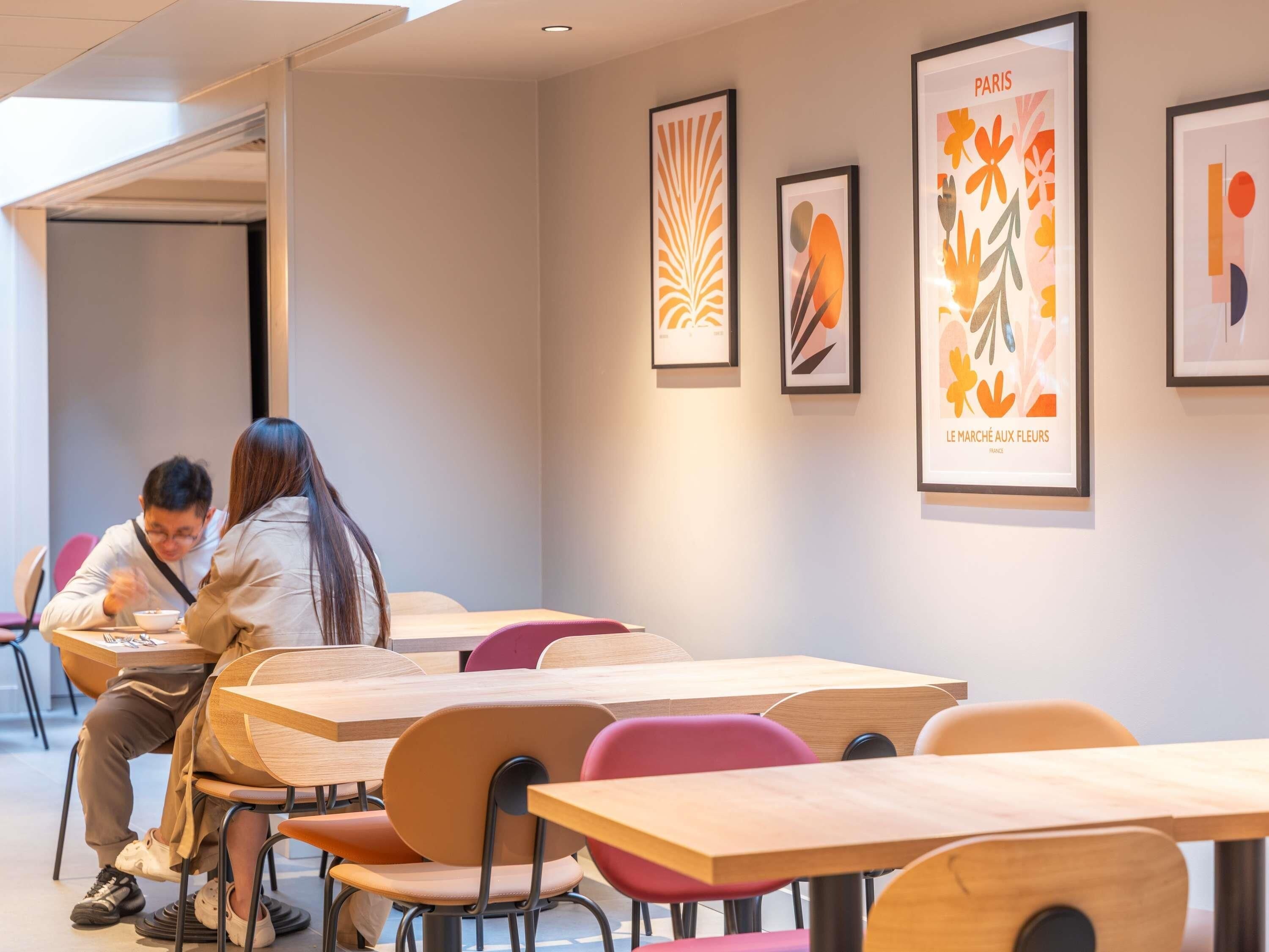 Un espace de restauration moderne avec des tables et des chaises en bois, des œuvres d’art encadrées accrochées aux murs, et une personne qui prend son repas.