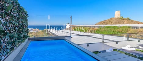 Piscina no terraço com vista para o mar e uma colina com uma construção.
