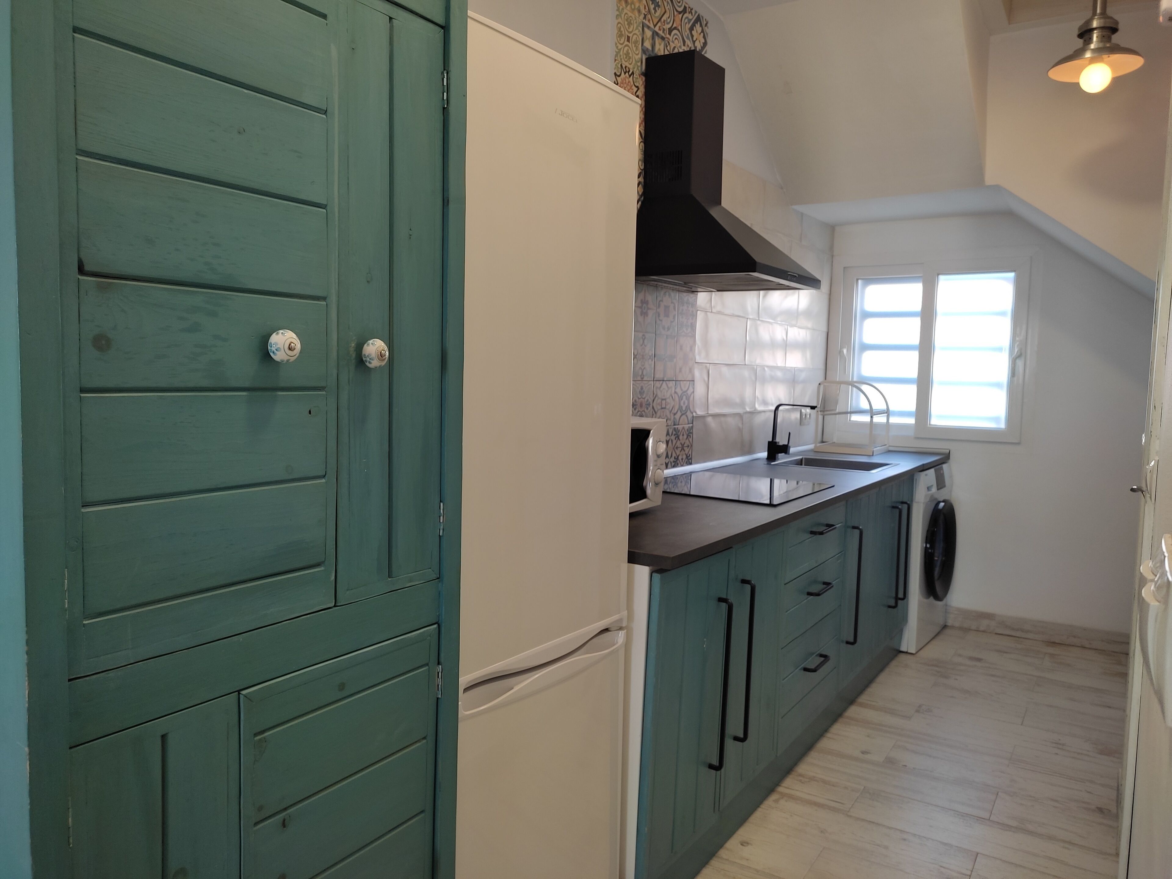 A kitchen with teal cabinets, a black range hood, a built-in oven, and a window above the sink.
