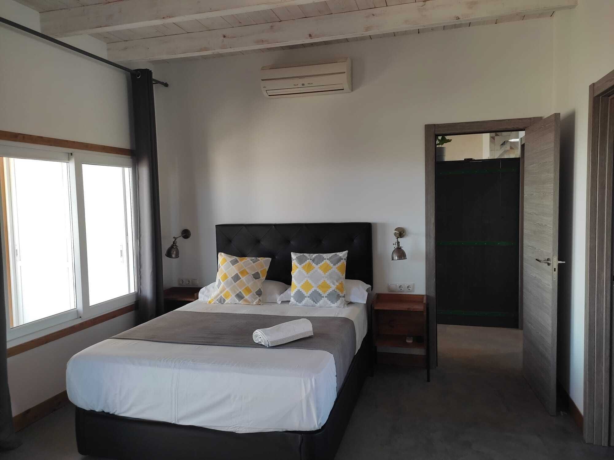 A bedroom with a bed, two nightstands, a wall-mounted air conditioner, and a door leading to another room.