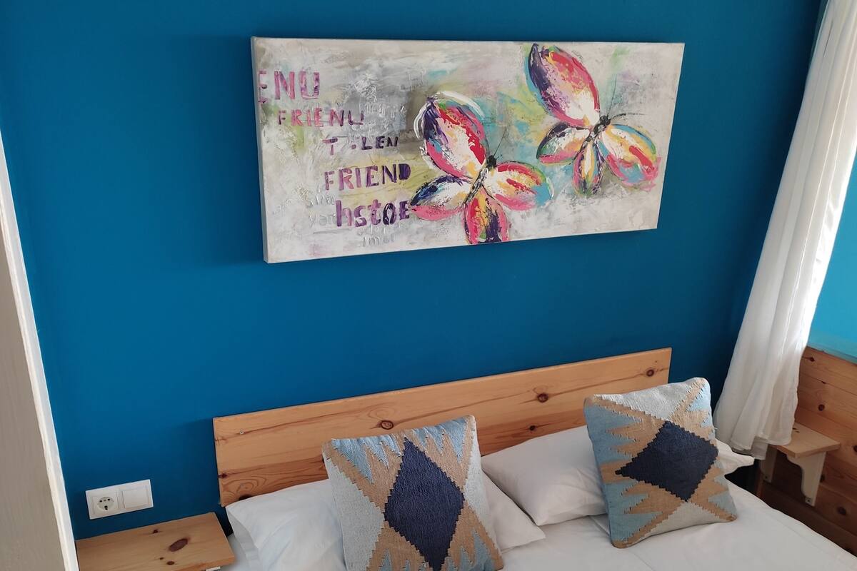 A bedroom with a blue wall, a wooden headboard, two pillows with geometric patterns, a painting of colorful flowers, and a white bedspread.