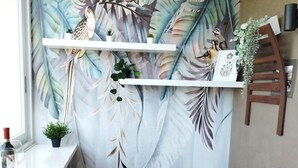 Una habitación acogedora con un sofá blanco, almohadones coloridos, una mesita roja y un mural de pájaros y hojas en la pared.