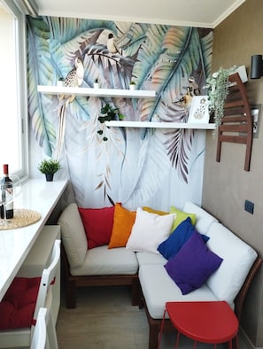 Una habitación acogedora con un sofá blanco, almohadones coloridos, una mesita roja y un mural de pájaros y hojas en la pared.