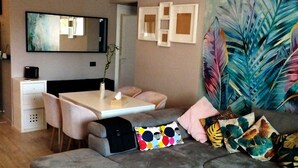 Una sala moderna con un sofá seccional gris, una mesa de comedor con sillas, un espejo grande y un mural vibrante con hojas tropicales en la pared.