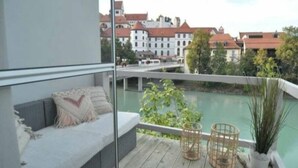 Een balkon met een bank, rieten manden en uitzicht op een rivier en gebouwen.