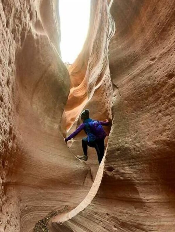 Une personne escalade un étroit canyon aux parois lisses et rougeâtres.