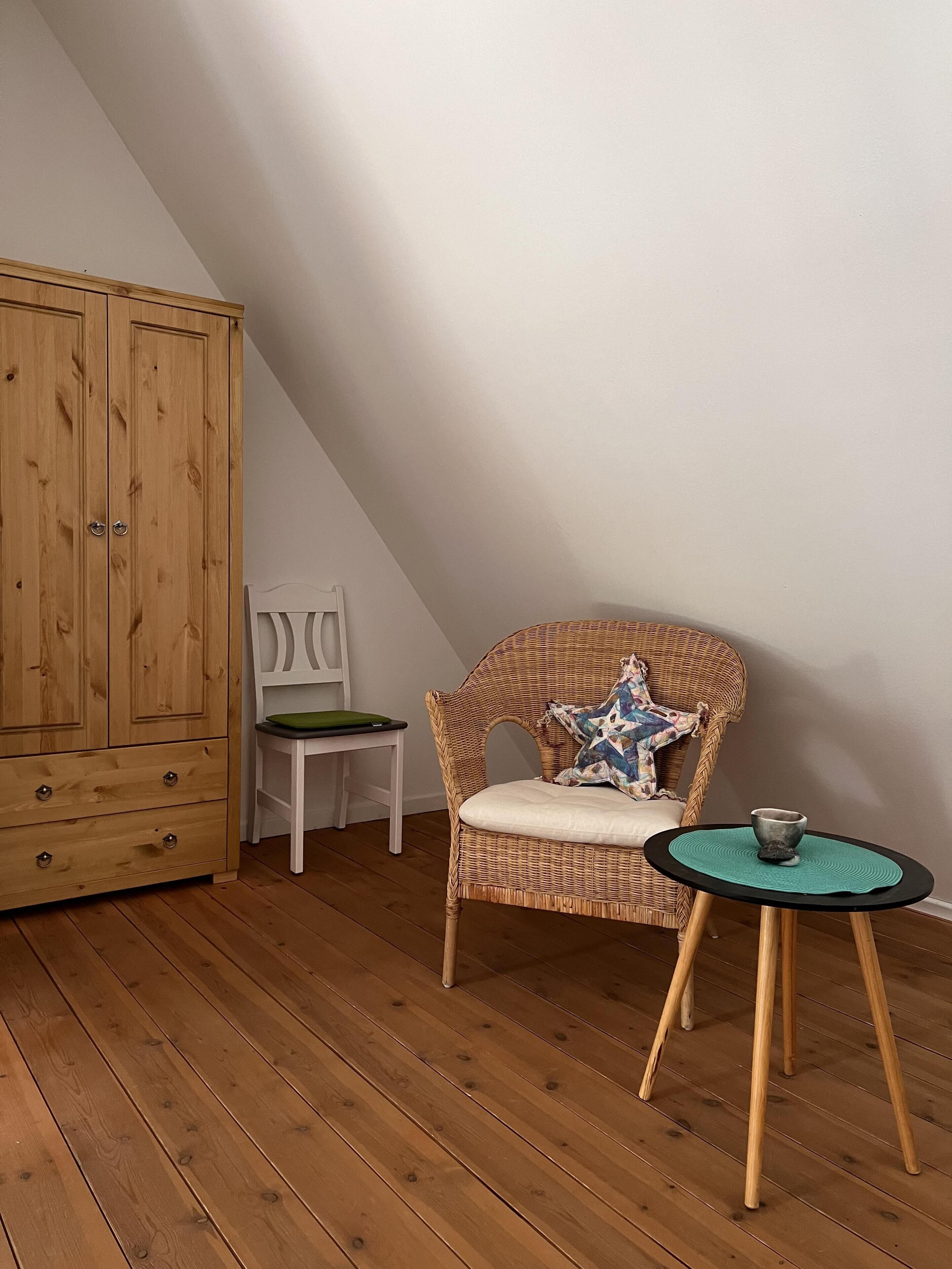 Una habitación con un armario de madera, una silla de mimbre con cojín, una mesita con una taza y una silla blanca.
