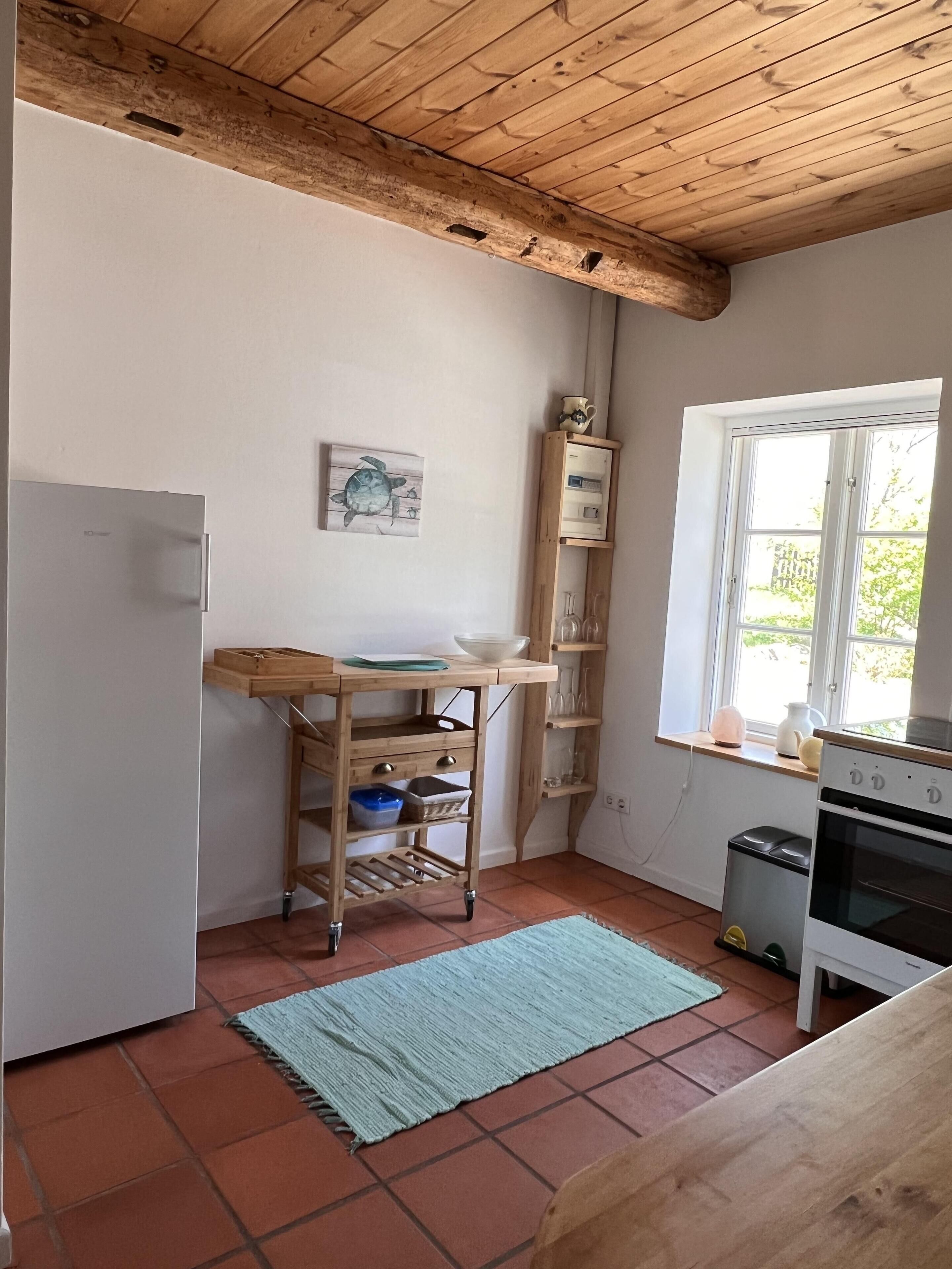A kitchen with a wooden ceiling, a window, a refrigerator, a wooden cart, and a tiled floor.
