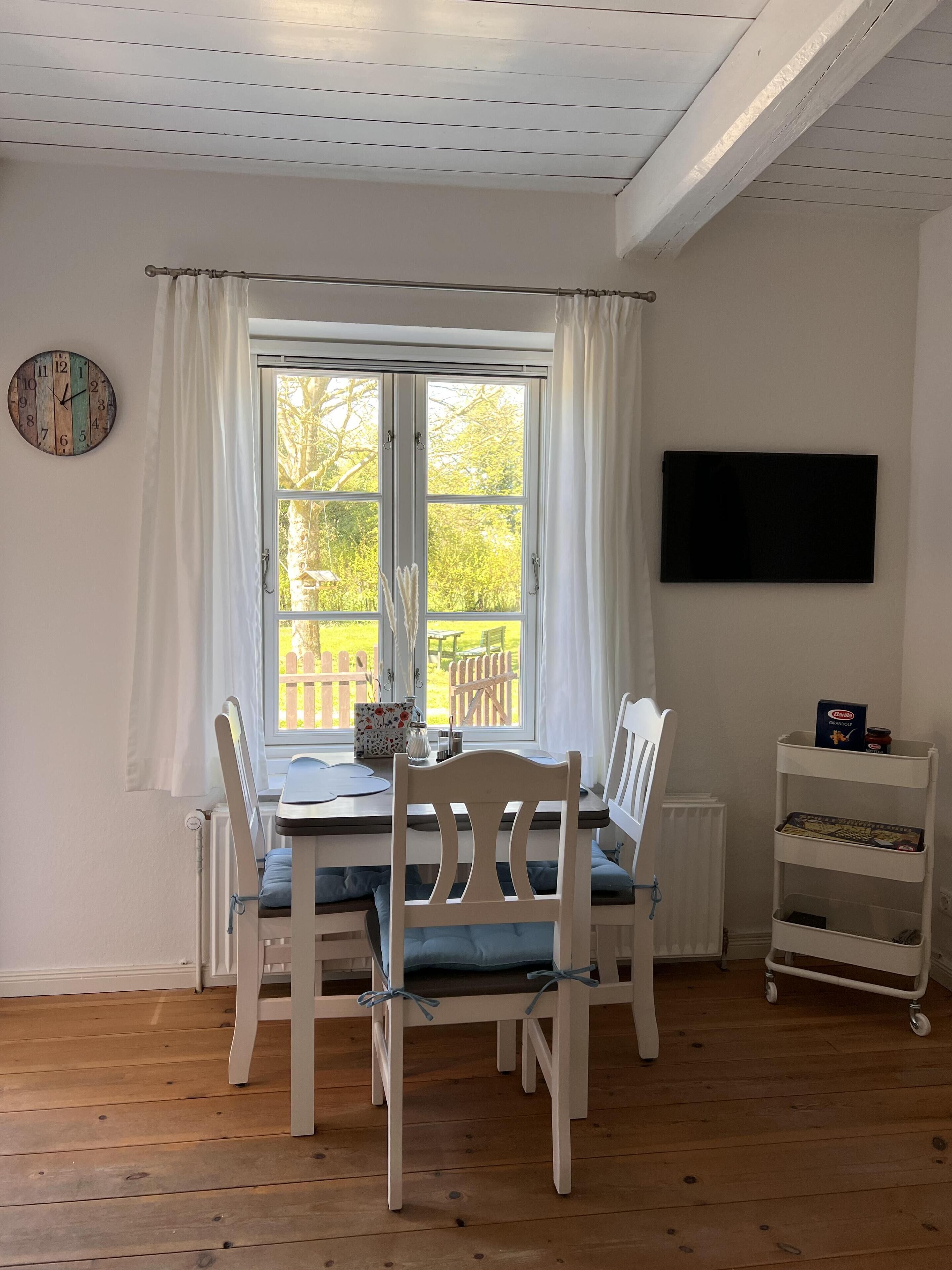 Un coin repas avec une table et des chaises, une horloge, une fenêtre avec des rideaux et un téléviseur fixé au mur.