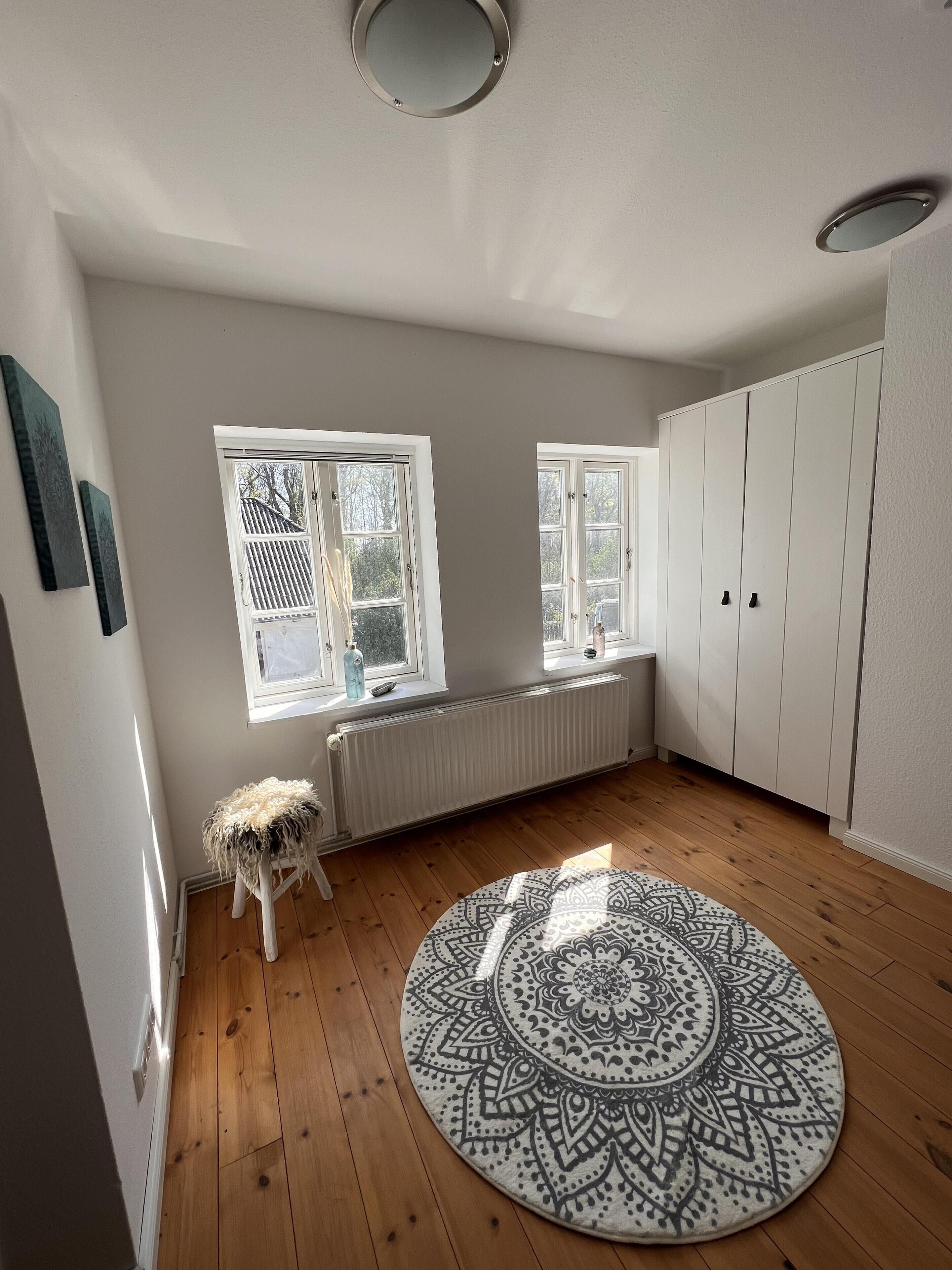 Ein Zimmer mit Holzboden, einem runden Teppich mit Muster, weißen Wänden, zwei Fenstern mit Jalousien, einem Heizkörper und einem weißen Kleiderschrank.