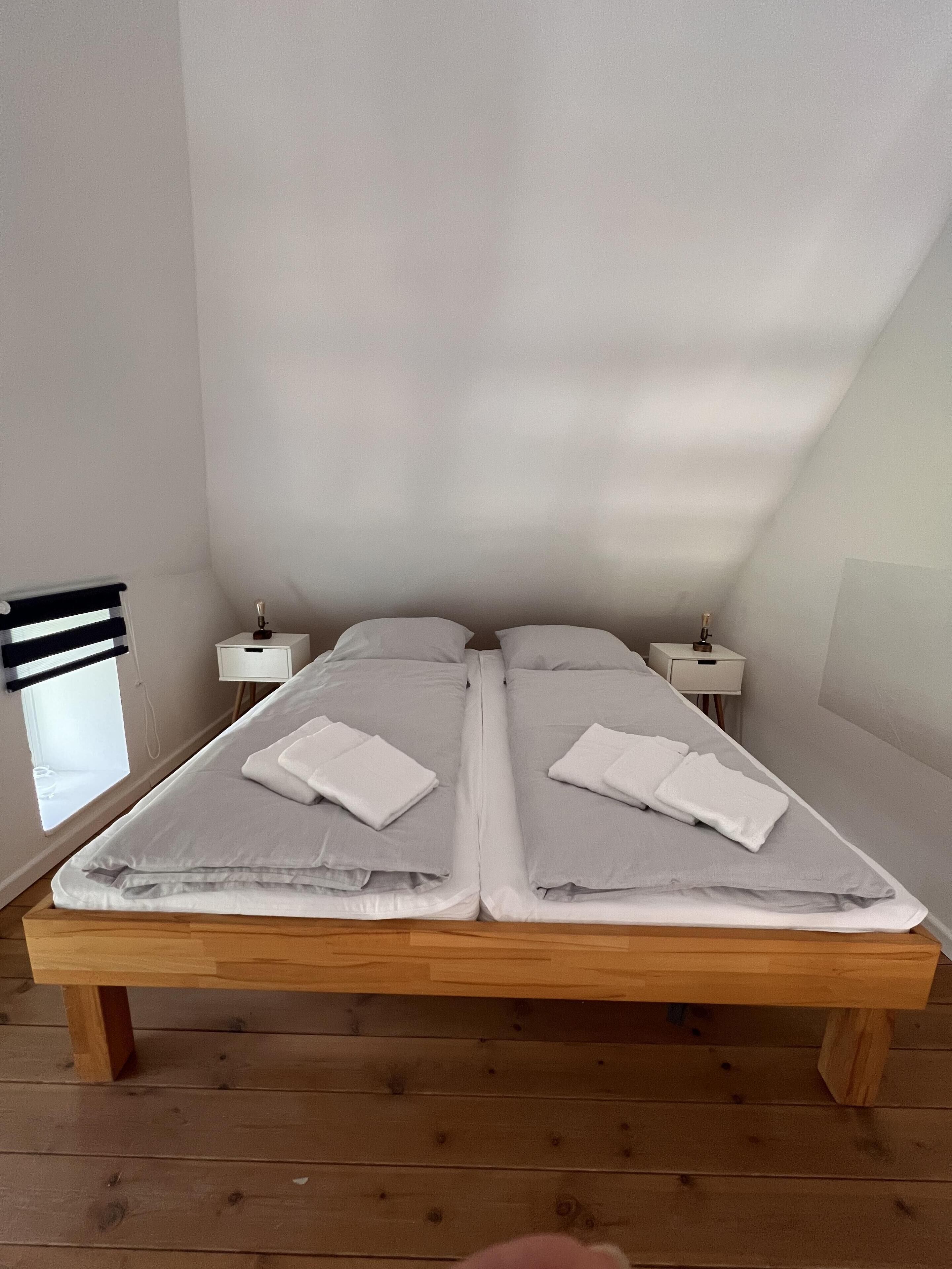 Ein Doppelbett mit weißen Leinen und Handtüchern, zwei Nachttische, ein Fenster mit Jalousien und ein Holzboden.