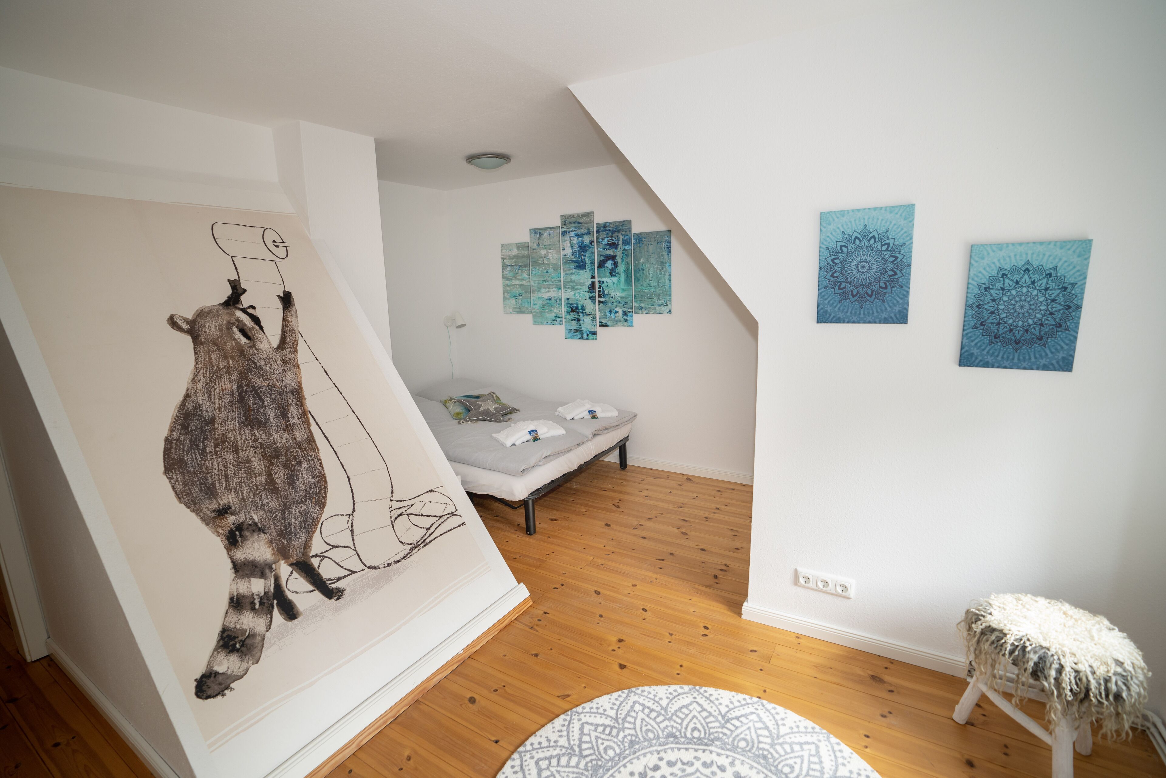Une pièce avec un grand mur décoré d’une fresque, un lit, un petit tabouret et trois œuvres d’art encadrées.
