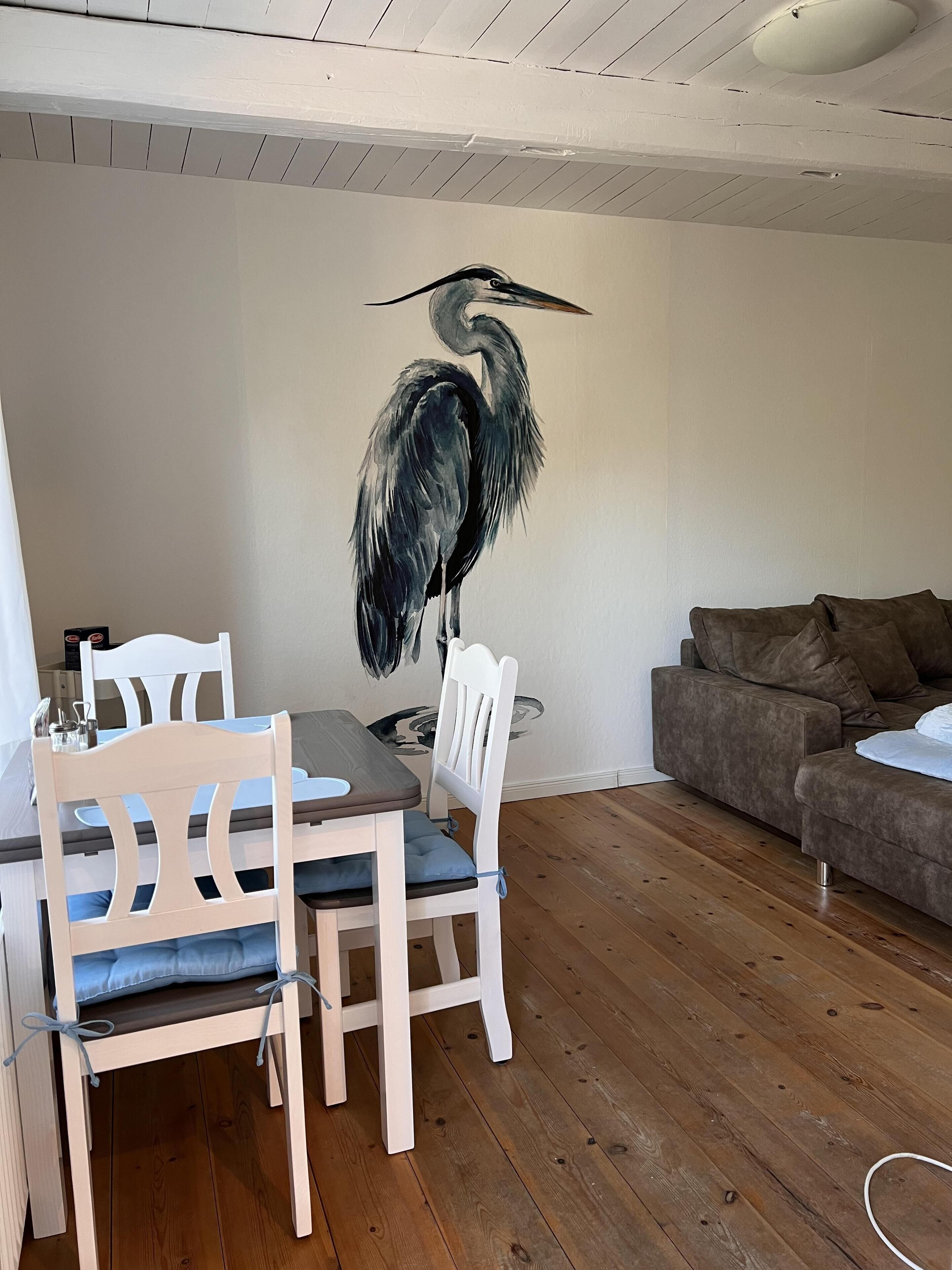 Ein Essbereich mit einem Holztisch und Stühlen, einer weißen Wand mit einem Vogelgemälde und einem braunen Ledersofa.