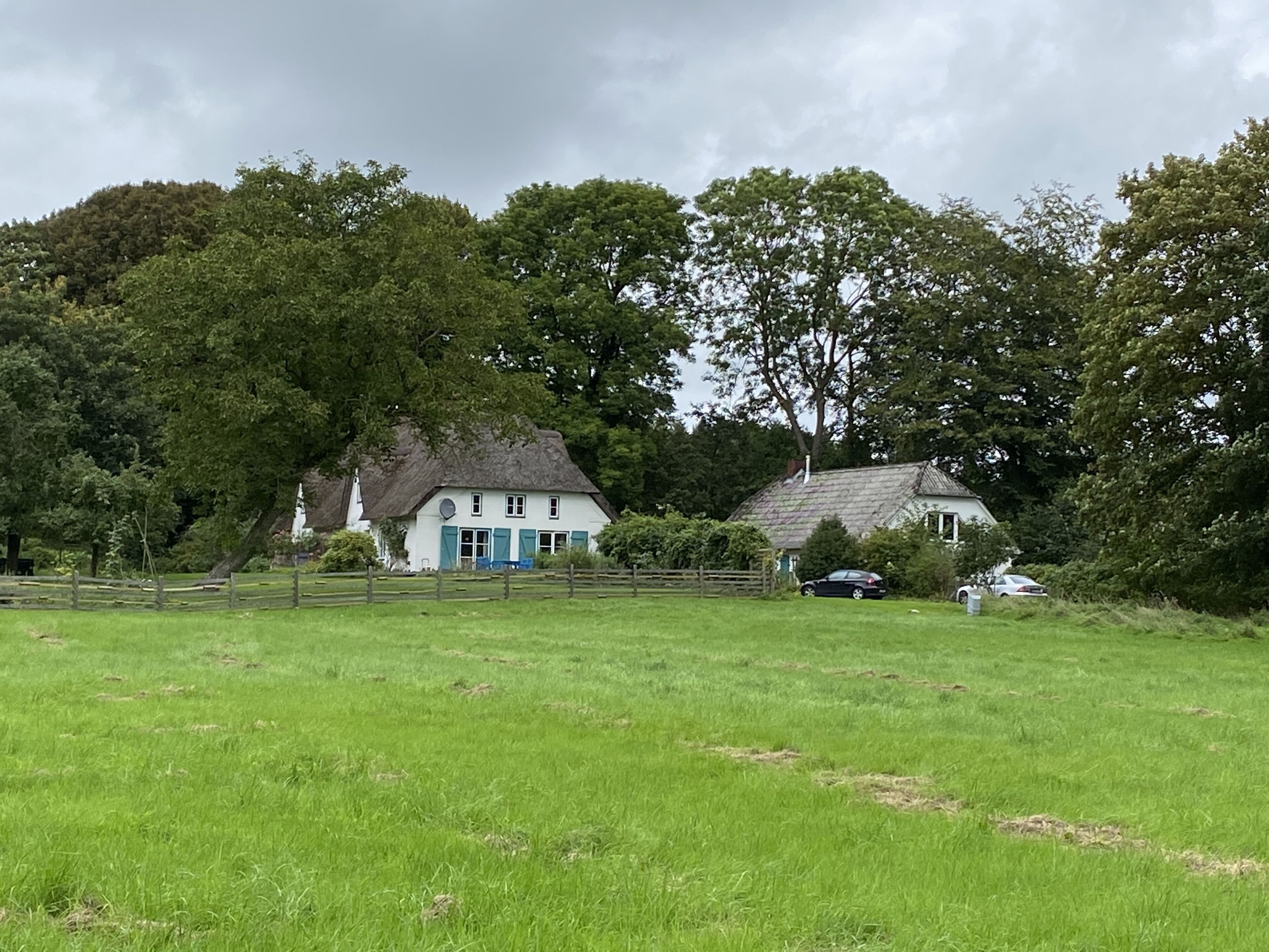 Un paysage rural avec une maison blanche aux volets bleus, entourée d’une végétation luxuriante et d’une construction au toit de chaume.
