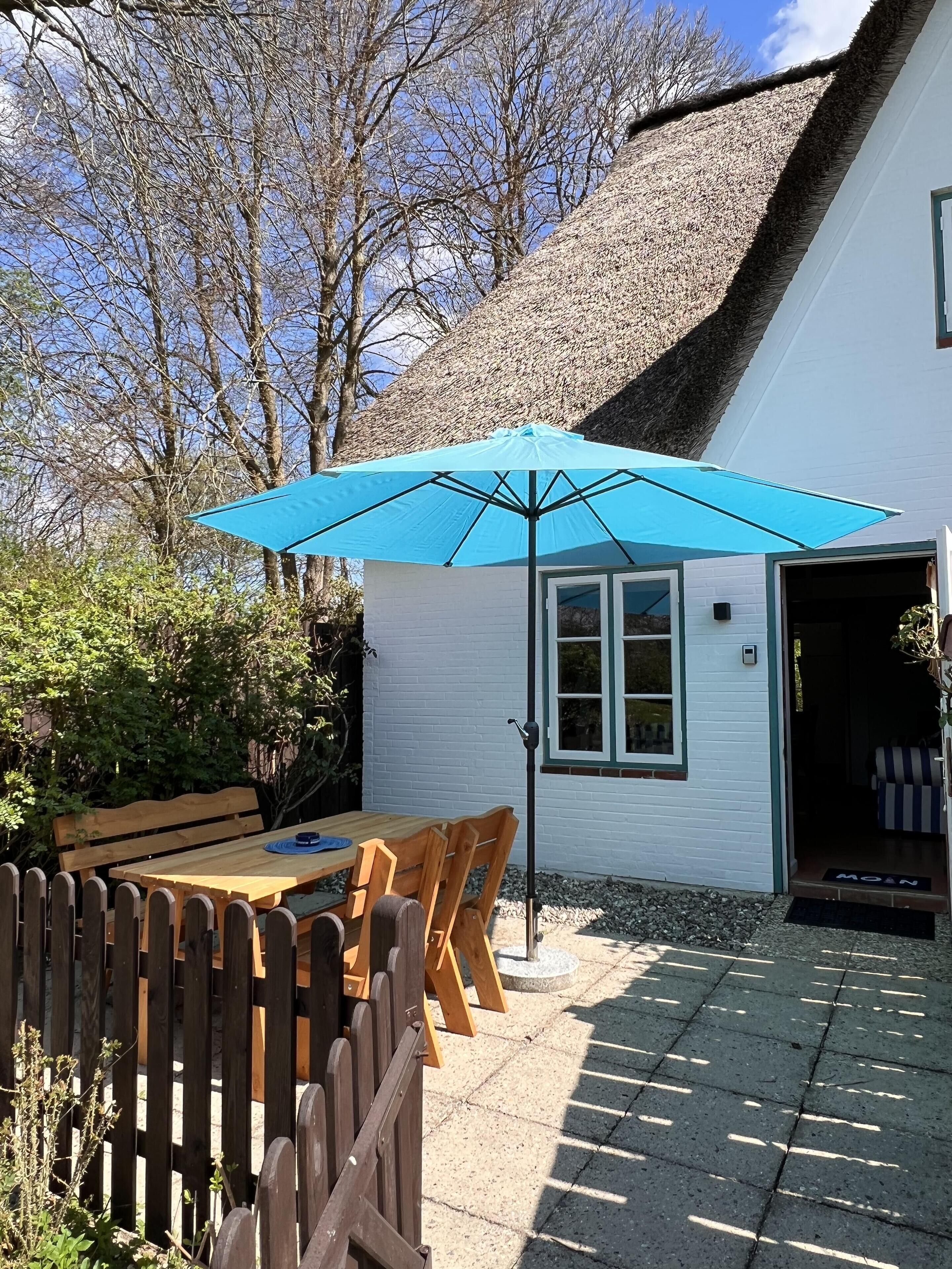 A wooden table and chairs under a blue umbrella, a white building with a thatched roof, and a wooden fence.