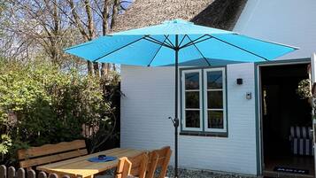 A wooden table and chairs under a blue umbrella, a white building with a thatched roof, and a wooden fence.