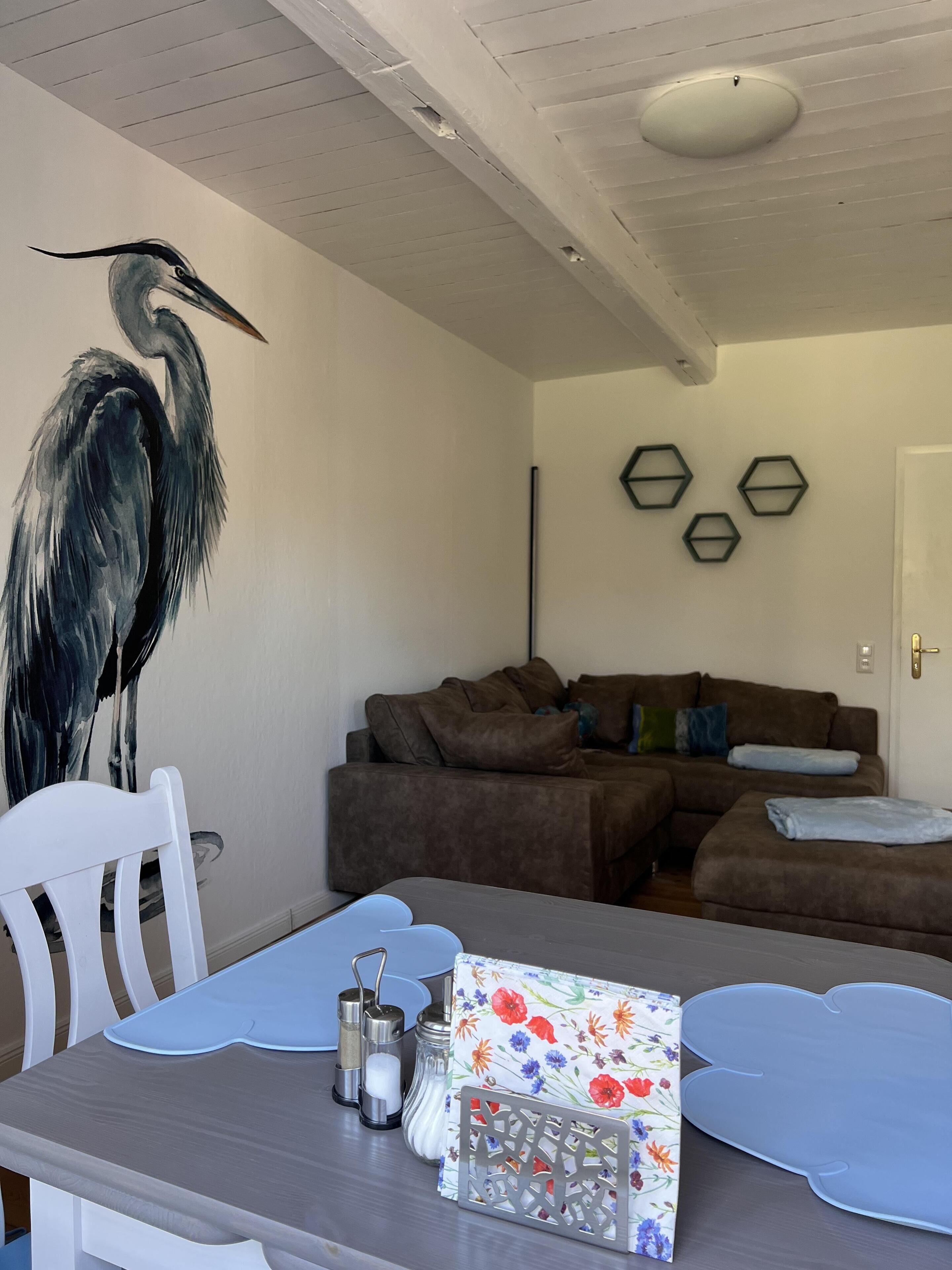 Ein Wohnzimmer mit einem großen Gemälde eines Vogels, einem braunen Ecksofa, einem weißen Sessel, einem Esstisch mit blauen Platzsets und sechseckigen Wanddekorationen.
