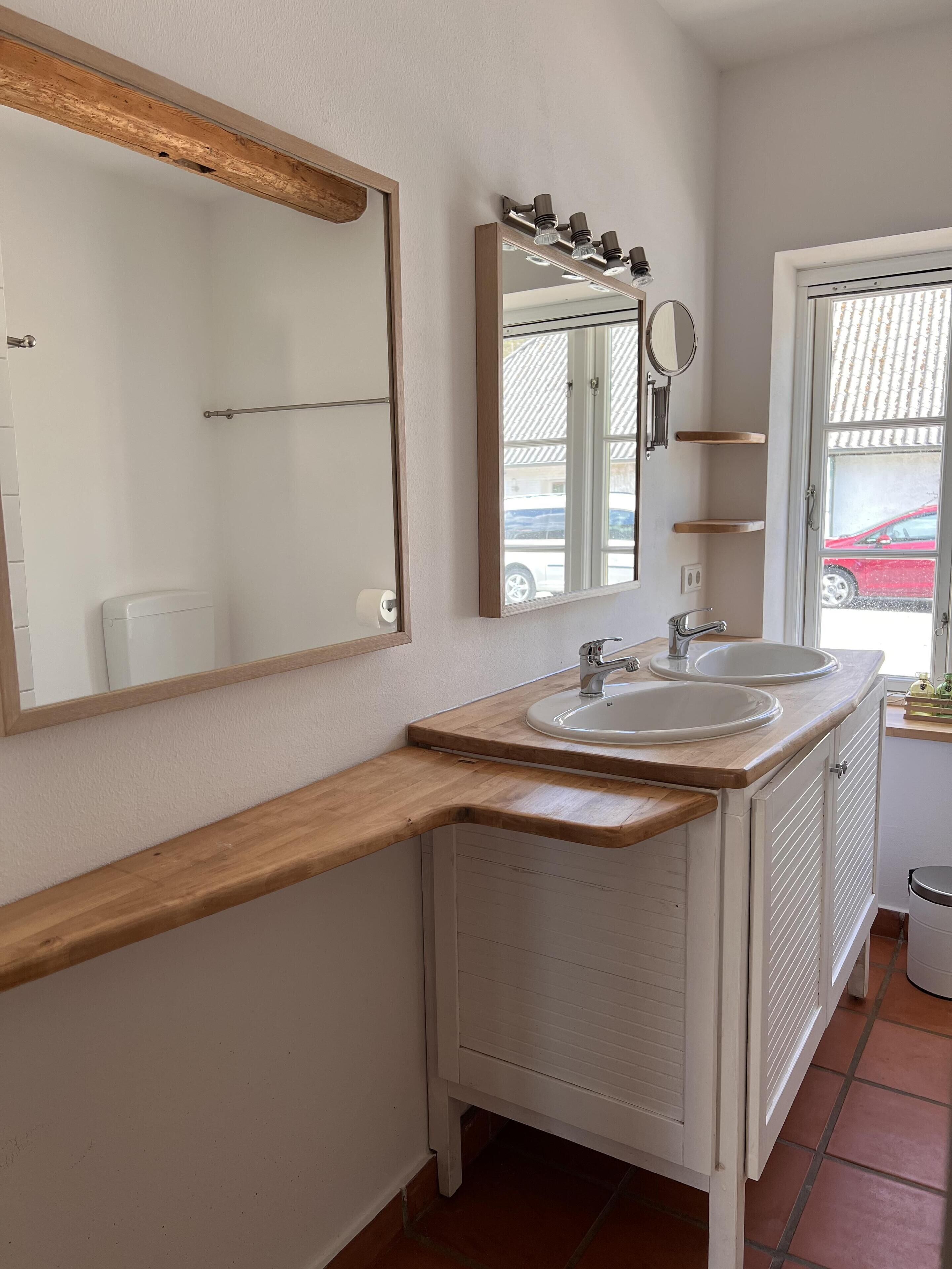 A bathroom with a wooden vanity, two sinks, a mirror, and a window with a view of a red car.