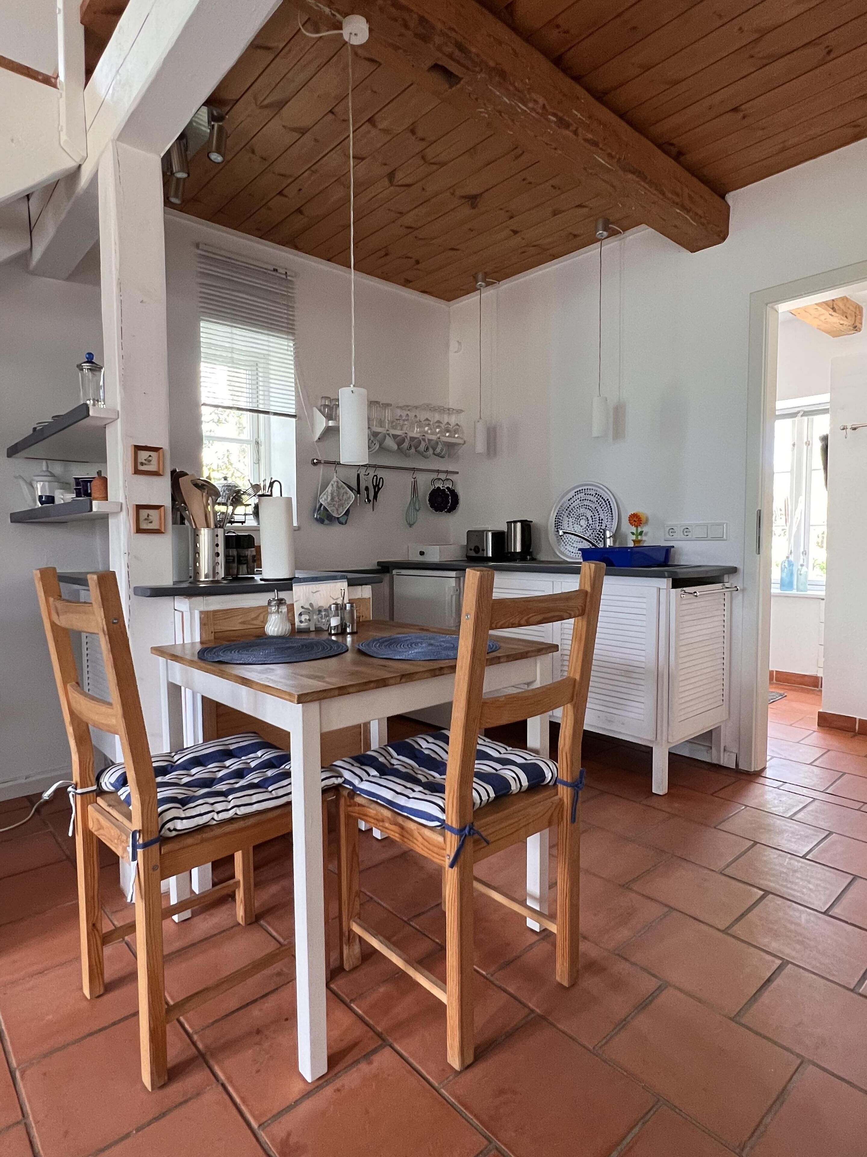 Uma sala de jantar com mesa e cadeiras de madeira, uma cozinha ao fundo e piso de azulejos.