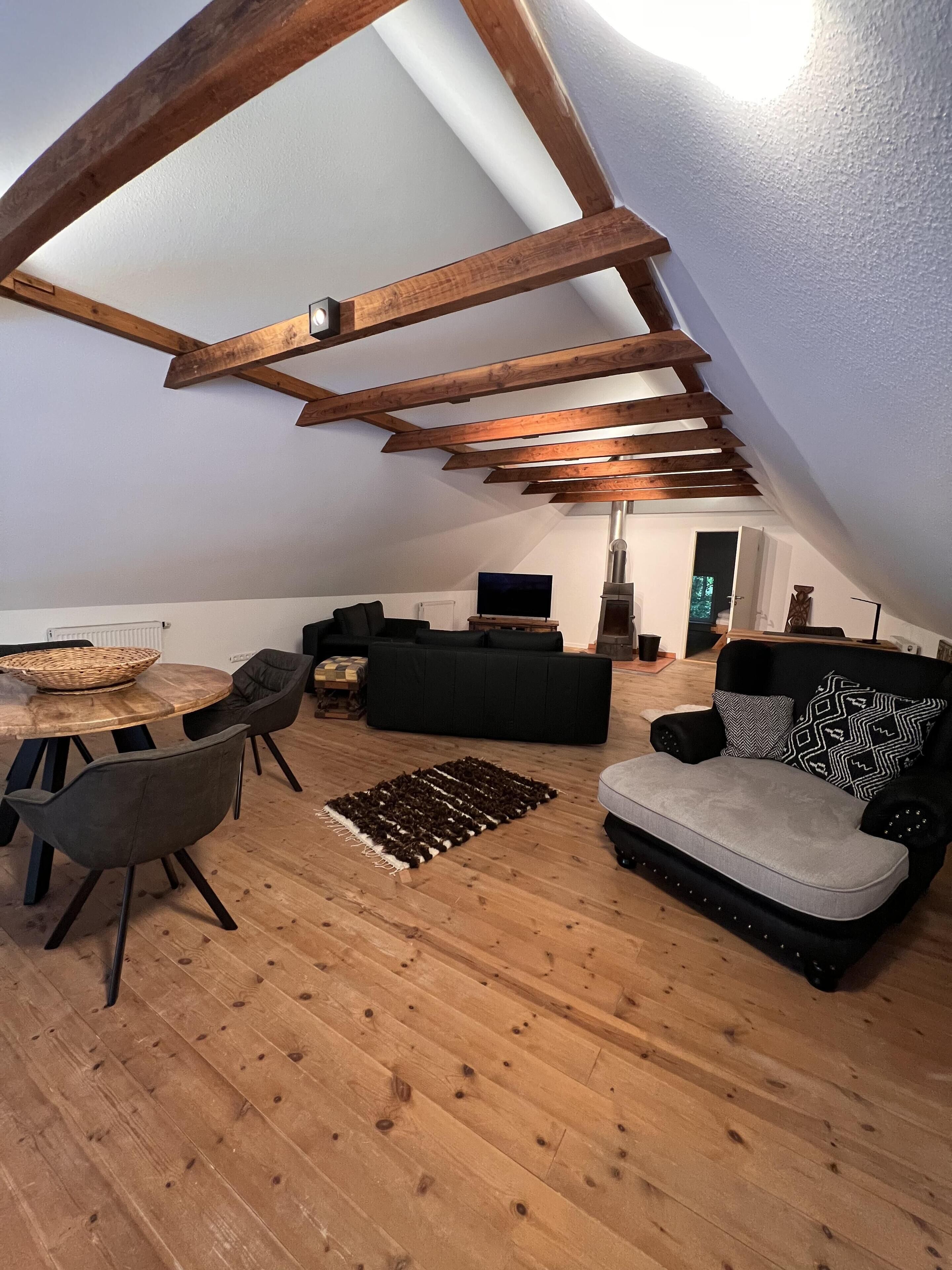 A loft-style living space with exposed wooden beams, a black sofa, a grey sofa, a dining table with chairs, and a television.