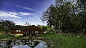 Une cabane en bois avec un étang, entourée d’arbres et bordée d’une pelouse.