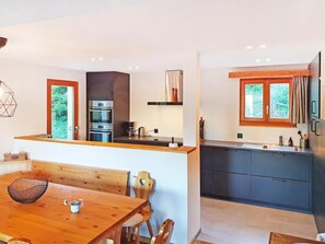 A modern kitchen with a wooden dining table and chairs, built-in appliances, and a large window with a view of greenery.