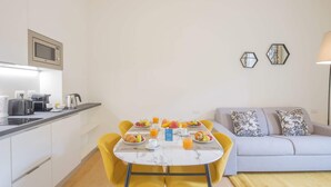 A compact kitchen with a microwave, toaster, and kettle, a dining table with breakfast items, and a sofa with patterned cushions. - Sweett | Fancy Duomo I (Milano)