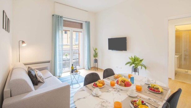 A modern living room with a sofa, a dining table set for breakfast, a TV mounted on the wall, and a balcony with a view.