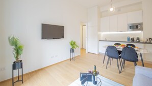 A modern apartment with a flat-screen TV mounted on the wall, a dining area with a table and chairs, a kitchen with a microwave, and a small potted plant.