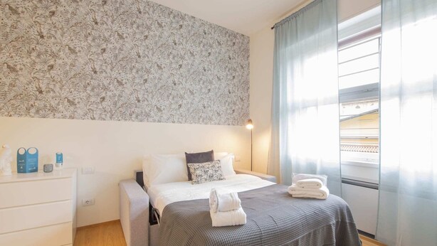 Une chambre avec un papier peint à motifs, un lit recouvert d’une couverture grise, une table de chevet avec une lampe et une fenêtre avec des rideaux.