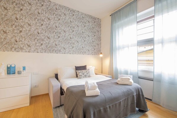 A bedroom with a patterned wallpaper, a bed with a grey bedspread, a bedside table with a lamp, and a window with curtains.