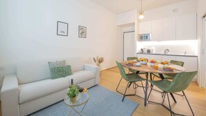 Una sala moderna con sofá, mesa de centro y comedor con sillas. Se observa una cocina al fondo, con microondas y fregadero.