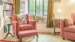 A cozy room with a red armchair, a wooden bookshelf, a window with curtains, and a small table with a lamp. - Saint Pancras Chapel (Watchet)