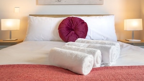 Un lit bien fait, avec du linge de lit blanc, un plaid rouge et deux serviettes blanches enroulées. On y trouve deux lampes de chevet, une carte du monde accrochée au mur et une tête de lit en bois.