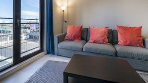 Un salon moderne avec un canapé gris, trois coussins orange, une table basse noire et une grande fenêtre donnant sur la ville.