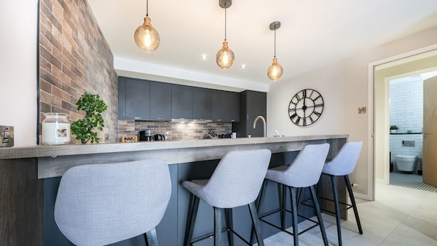 Une cuisine moderne avec un mur d’accent en briques, un îlot central doté de tabourets de bar, un éclairage suspendu et une grande horloge murale.